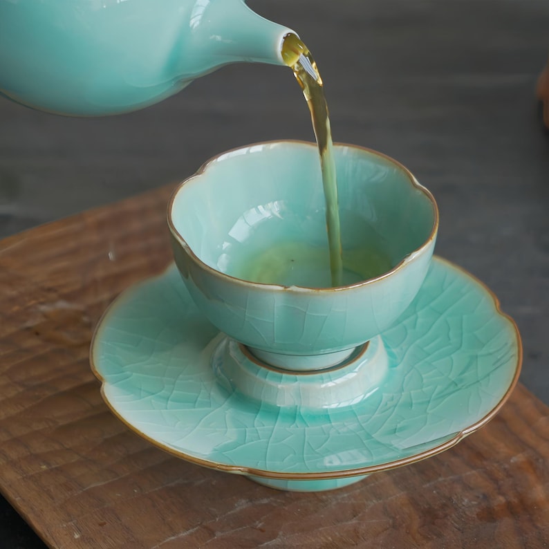 May include: A light mint green ceramic tea cup and saucer set with a delicate crackle glaze finish and gold-toned rims. A matching teapot pours golden tea into the cup, which features a scalloped edge design on both the cup and saucer.