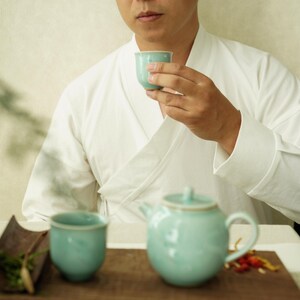 May include: A person holding a light blue ceramic teacup. A matching teapot and teacup sit on a wooden tray. The person is wearing a white robe. The tea set is a soft, muted blue-green colour.