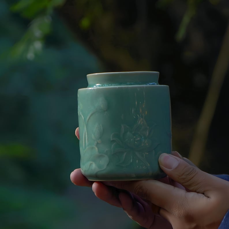 May include: A teal ceramic jar with a lotus flower design. The jar is cylindrical with a slightly flared rim. The lotus flower and leaves are carved into the surface of the jar. The jar is held in a person's hand.