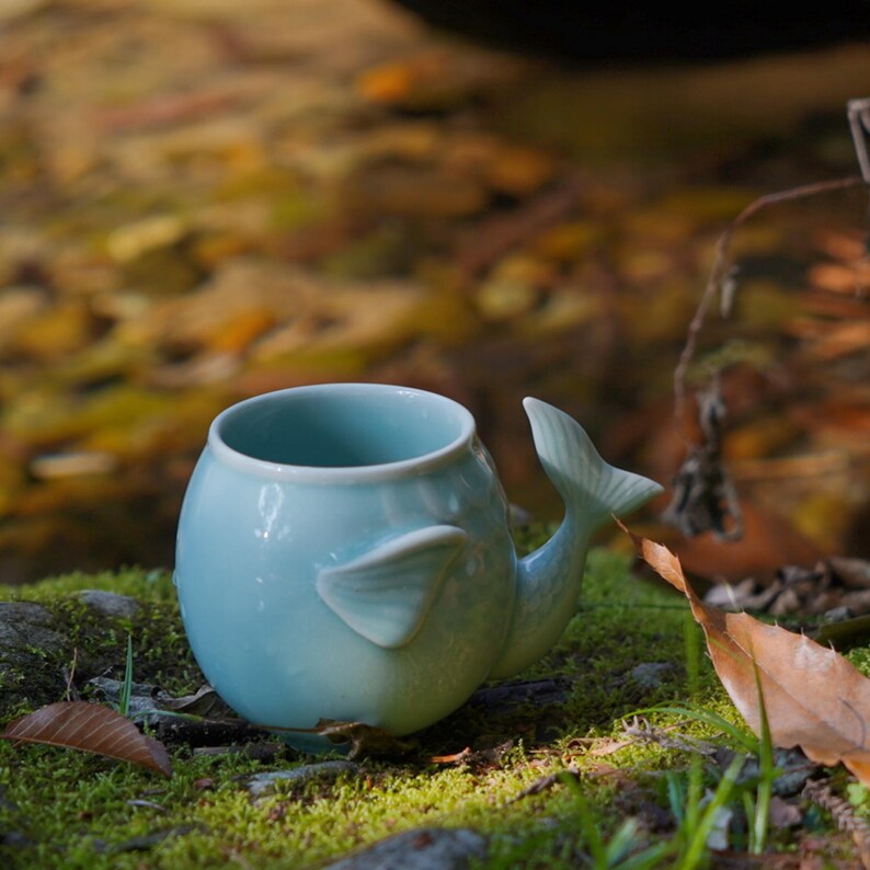 May include: A light blue ceramic whale-shaped mug with a tail handle and fin details. The mug is sitting on a bed of green moss and leaves, creating a natural setting. The mug's design is whimsical and unique.
