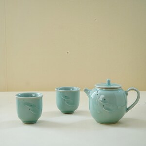 May include: A light teal ceramic tea set comprising a teapot with a lid and two teacups. Each piece features a delicate embossed design. The teapot has a rounded body, a spout, and a handle. The teacups are small and cylindrical.