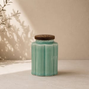 May include: A turquoise ceramic jar with a ribbed design and a dark wood lid. The jar has a rounded base and a cylindrical body. The lid is flat and circular. The jar is sitting on a light-colored surface.