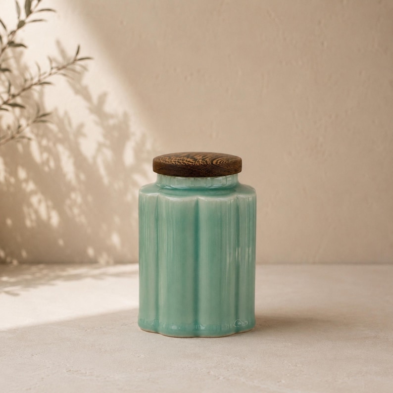 May include: A turquoise ceramic jar with a ribbed design and a dark wood lid. The jar has a rounded base and a cylindrical body. The lid is flat and circular. The jar is sitting on a light-colored surface.