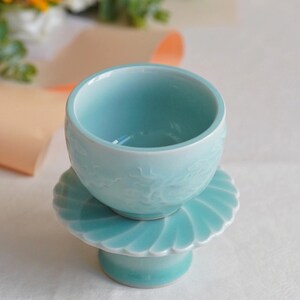 May include: A light blue ceramic teacup with a matching saucer and pedestal. The teacup has a rounded shape and a detailed dragon design. The saucer and pedestal have a scalloped edge design. The set is a soft, pastel blue color.