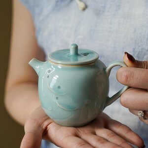 May include: A pale turquoise ceramic teapot with a lid and handle. The teapot has a rounded body and is decorated with embossed fish designs. The spout and handle are curved. The teapot is held in a person's hands.