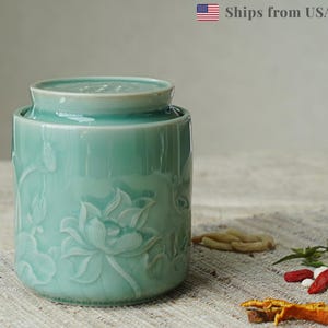 May include: A light teal ceramic jar with a lid, featuring embossed lotus flower designs. The jar is cylindrical and sits on a textured surface. The jar is approximately 6 inches tall.