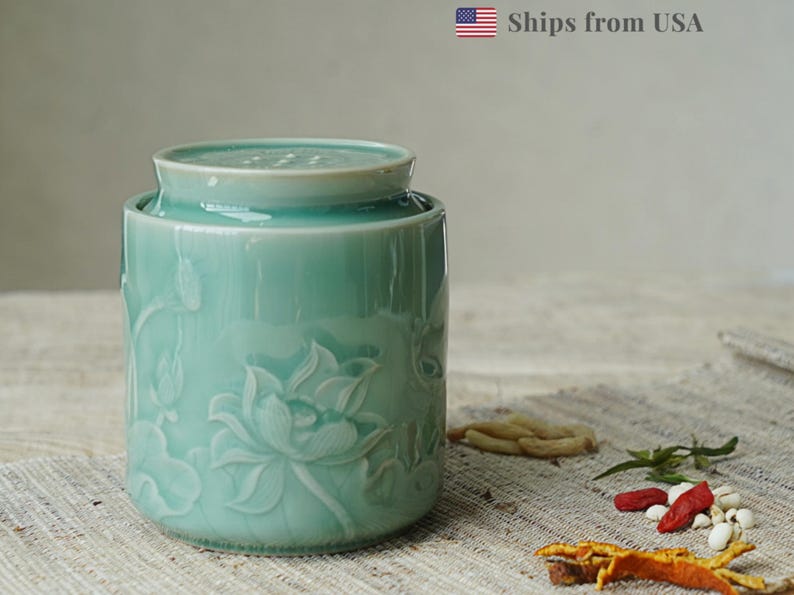 May include: A light teal ceramic jar with a lid, featuring embossed lotus flower designs. The jar is cylindrical and sits on a textured surface. The jar is approximately 6 inches tall.