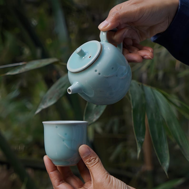 May include: A pale blue ceramic teapot with a matching lid and spout is being poured into a pale blue teacup. Both have a speckled design. The background features blurred green foliage. The teapot and cup are likely for tea.