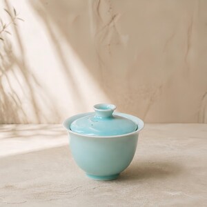 K&ouml;nnte beinhalten: Eine hellblaue Gaiwan-Teeschale aus Keramik mit passendem Deckel. Die Schale hat eine glatte, gl&auml;nzende Oberfl&auml;che und einen ausgestellten Rand, w&auml;hrend der Deckel einen kleinen, erh&ouml;hten Knopfgriff in der Mitte besitzt.