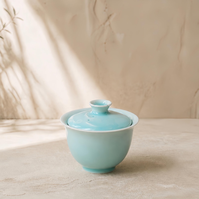 K&ouml;nnte beinhalten: Eine hellblaue Gaiwan-Teeschale aus Keramik mit passendem Deckel. Die Schale hat eine glatte, gl&auml;nzende Oberfl&auml;che und einen ausgestellten Rand, w&auml;hrend der Deckel einen kleinen, erh&ouml;hten Knopfgriff in der Mitte besitzt.