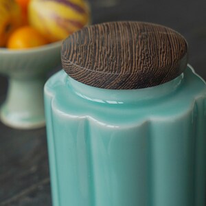 May include: A turquoise ceramic jar with a wooden lid. The jar has a fluted design and a glossy finish. The lid is round with a wood grain pattern. A bowl of fruit is visible in the background.