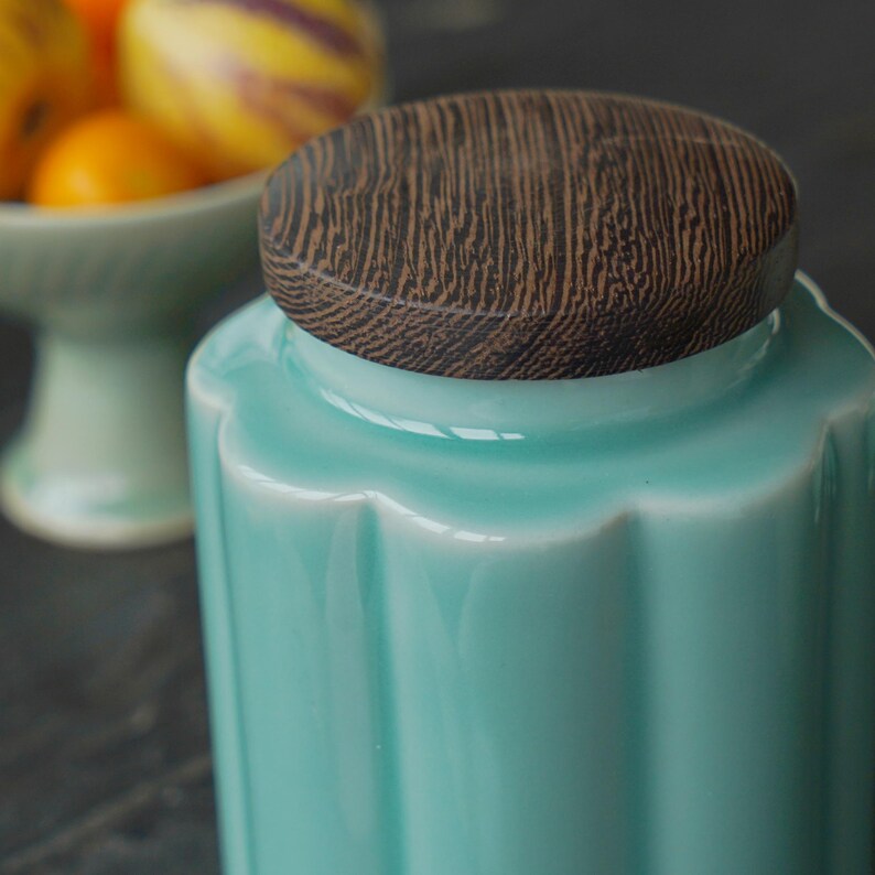 May include: A turquoise ceramic jar with a wooden lid. The jar has a fluted design and a glossy finish. The lid is round with a wood grain pattern. A bowl of fruit is visible in the background.