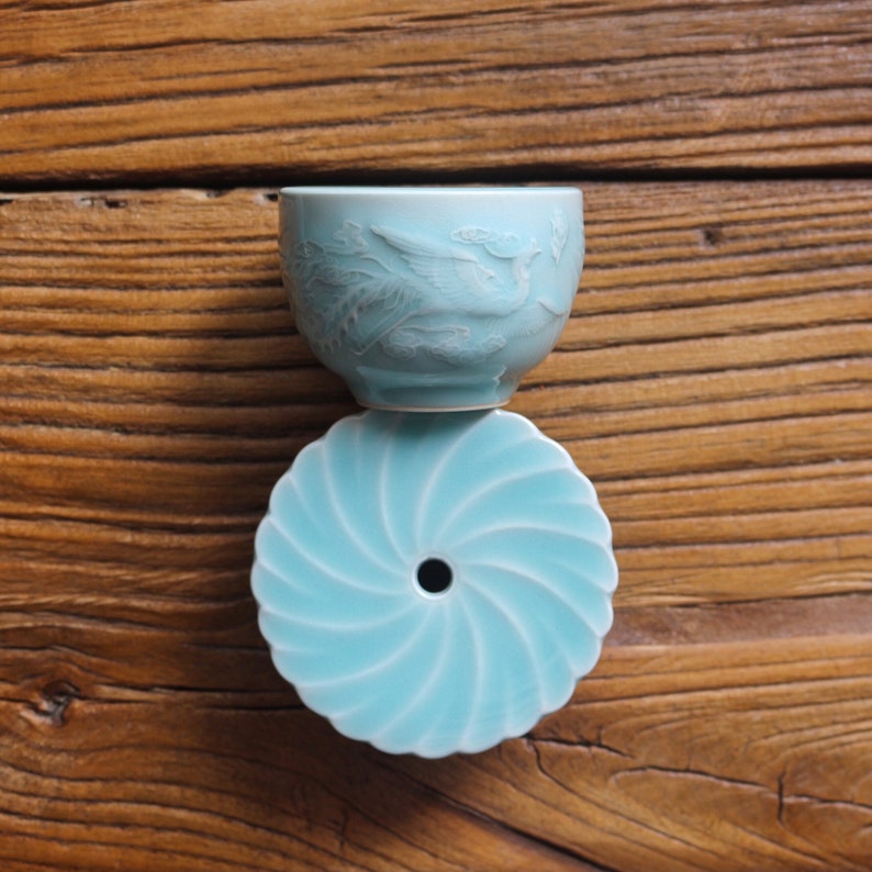 May include: A light blue ceramic teacup with a detailed relief of a bird and clouds, resting on a matching saucer. The saucer has a spiral design with a central hole. The teacup and saucer are set against a wooden background.