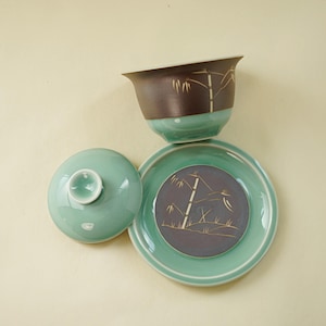 May include: A green ceramic teacup with a lid and saucer. The cup and saucer have a brown and purple glaze with a gold bamboo design.