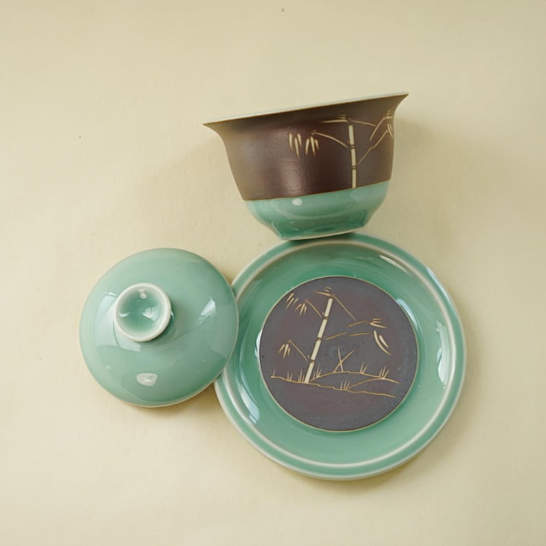 May include: A green ceramic teacup with a lid and saucer. The cup and saucer have a brown and purple glaze with a gold bamboo design.