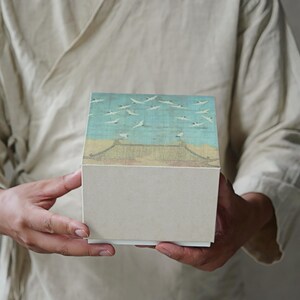May include: A square box with a lid featuring a vintage-style illustration of flying birds over a building. The box has a light beige base and a lid with a blue, yellow, and white design. The box is held by a person.