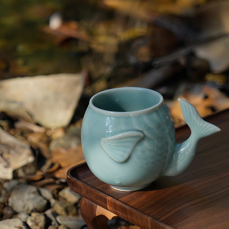 May include: A light blue ceramic mug with a fish tail handle and a fish fin detail. The mug is sitting on a wooden surface. The mug has a unique design and is likely used for drinking beverages.