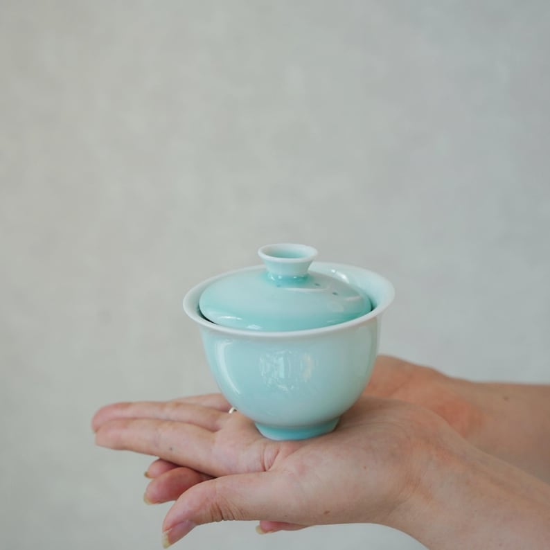 K&ouml;nnte beinhalten: Eine hellblaue Gaiwan-Teeschale aus Keramik mit passendem Deckel. Die Schale hat eine glatte, gl&auml;nzende Oberfl&auml;che und einen kleinen, ausgestellten Knopf in der Mitte des Deckels. Das Gef&auml;&szlig; hat einen abgerundeten K&ouml;rper, der sich zu einem kleinen Fu&szlig; verj&uuml;ngt.
