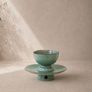 May include: A teal ceramic bowl with a scalloped rim sits atop a matching pedestal base. The base has a round, flat plate and a diamond-shaped cut-out. The edges are accented with a thin, brown trim. The bowl and base are on a neutral surface.