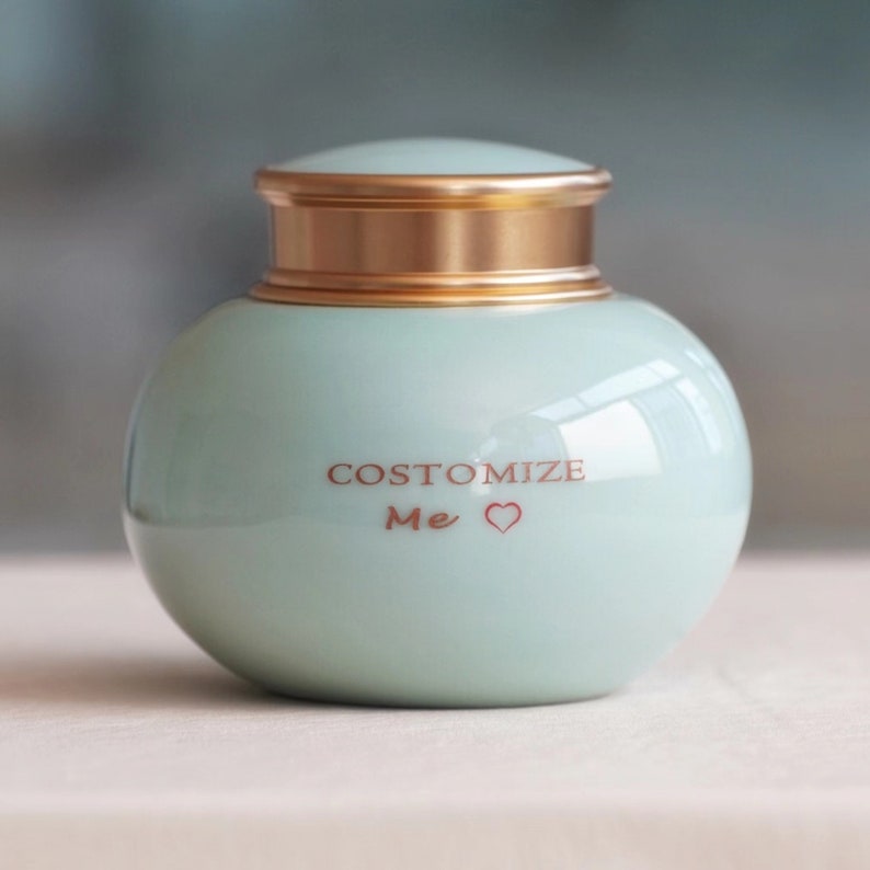 Puede incluir: Un tarro de cer&aacute;mica verde claro, de forma esf&eacute;rica, con tapa dorada. El tarro tiene la inscripci&oacute;n "COSTOMIZE Me" en rojo, con un s&iacute;mbolo de coraz&oacute;n. Probablemente es para decoraci&oacute;n o almacenamiento.