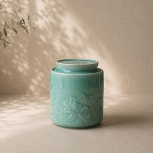 May include: A turquoise ceramic jar with a lid, featuring a detailed embossed lotus flower design. The jar has a cylindrical shape and is likely used for storage or decoration. The background is a neutral color.