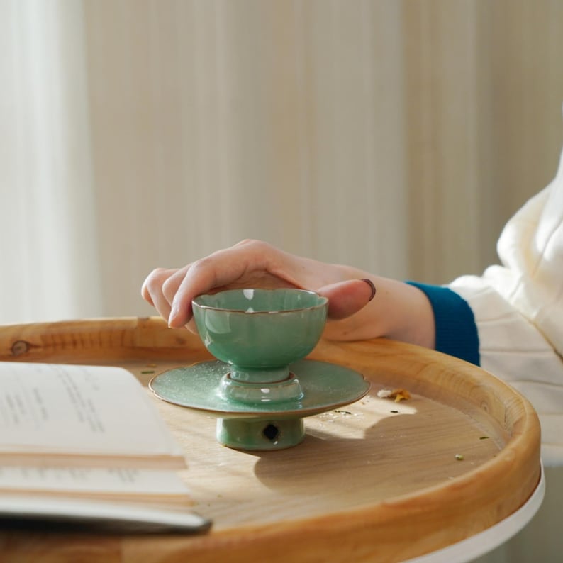 May include: A light green ceramic tea cup with a matching saucer, featuring a pedestal base and a scalloped rim. The glaze has a subtle speckled texture with a brown edge along the rim of the cup and the saucer, creating a cohesive look.