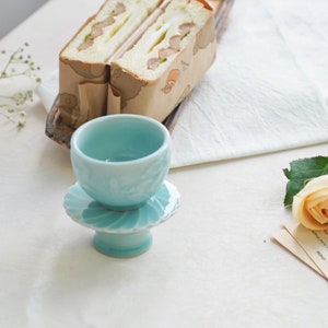 May include: A light blue ceramic teacup and saucer set. The teacup sits atop a pedestal base with a scalloped edge. Two sandwiches wrapped in brown paper are in the background, along with a white cloth and a rose.