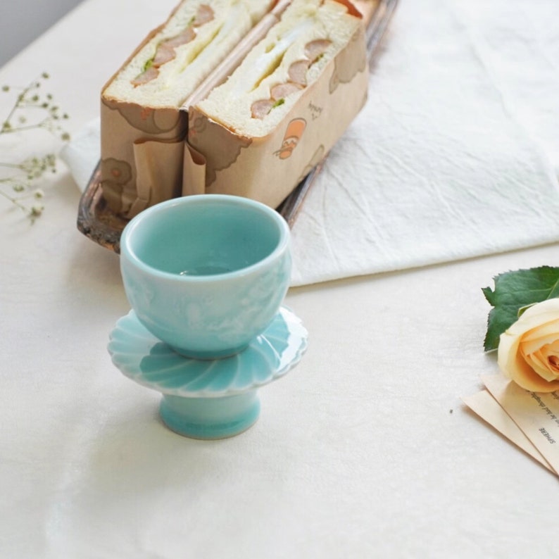 May include: A light blue ceramic teacup and saucer set. The teacup sits atop a pedestal base with a scalloped edge. Two sandwiches wrapped in brown paper are in the background, along with a white cloth and a rose.