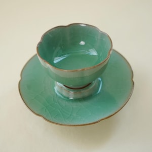 May include: A light green ceramic tea cup and matching saucer set featuring a scalloped edge design and a visible crackle glaze finish. The cup sits centered on the saucer, both displaying a consistent celadon hue with dark brown rims.