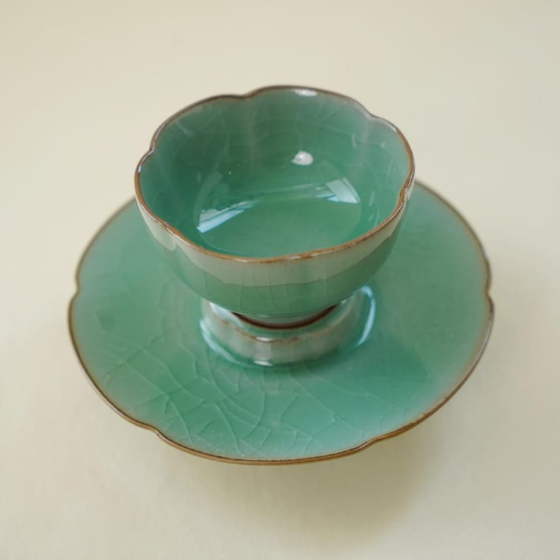 May include: A light green ceramic tea cup and matching saucer set featuring a scalloped edge design and a visible crackle glaze finish. The cup sits centered on the saucer, both displaying a consistent celadon hue with dark brown rims.
