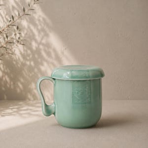 May include: A light green ceramic mug with a matching lid. The mug has a curved handle and a slightly textured surface. The lid sits flush on top. The mug is approximately 10 cm tall.