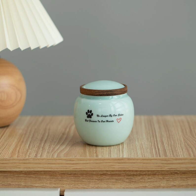 May include: A light blue ceramic pet urn with a wooden lid. The urn has a black paw print and the text "No Longer By Our Side But Forever In Our Hearts" with a red heart.