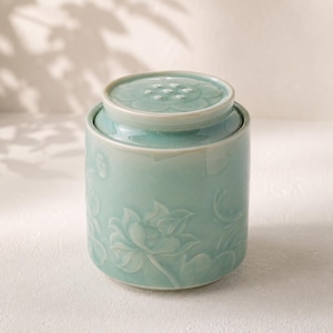 Può includere: Un barattolo cilindrico in ceramica verde chiaro con coperchio abbinato. L'esterno presenta un design in rilievo di fiori e foglie di loto. Il coperchio ha una parte superiore circolare con un motivo intagliato di baccello di loto.