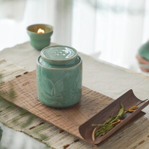 May include: A light teal ceramic tea caddy with a lid, placed on a wooden serving board. The caddy has a floral design. A small teal candle holder with a lit candle is in the background. A wooden tray with tea leaves is also present.