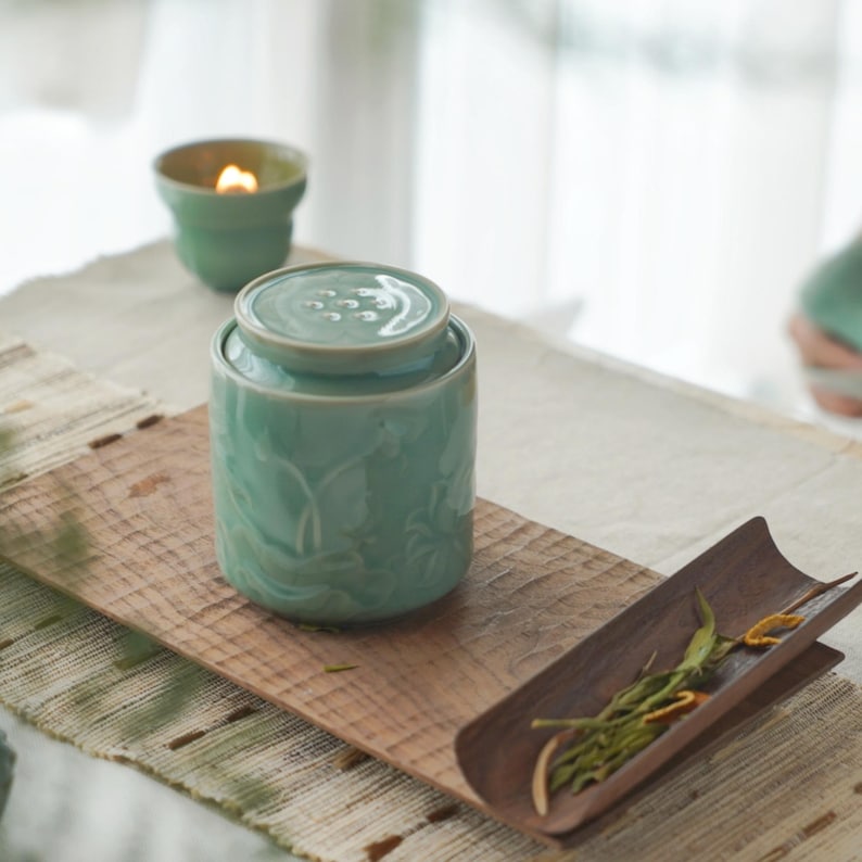 May include: A light teal ceramic tea caddy with a lid, placed on a wooden serving board. The caddy has a floral design. A small teal candle holder with a lit candle is in the background. A wooden tray with tea leaves is also present.