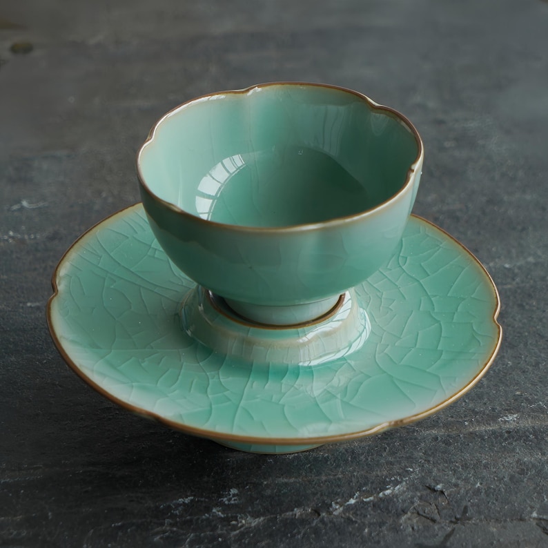 May include: A light mint green ceramic tea cup and matching saucer set featuring a delicate crackle glaze finish. The cup has a scalloped rim and a pedestal base, while the saucer has a raised center and a matching scalloped edge.