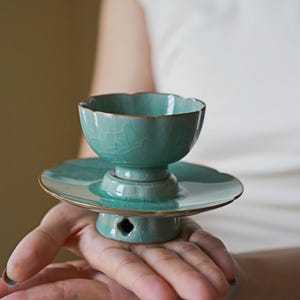 May include: A light blue-green ceramic tea cup with a scalloped rim and a matching saucer. The glaze features a distinct crackle pattern throughout the surface. The base of the saucer has a small, four-pointed cutout detail.