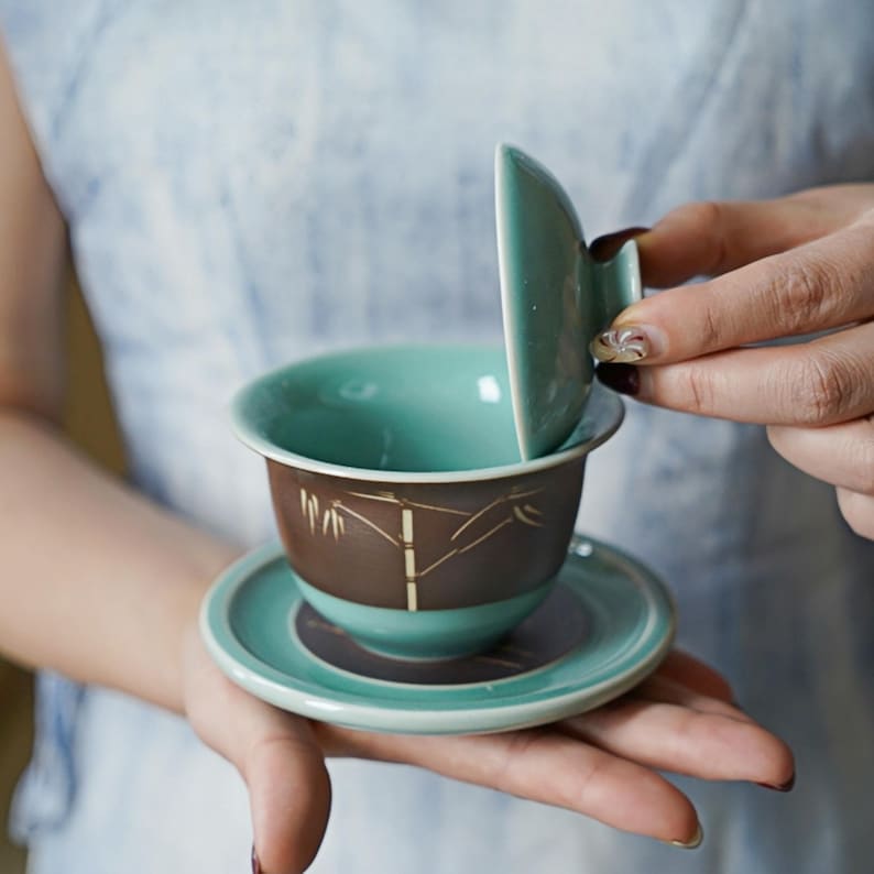 May include: A teal ceramic teacup with a brown and gold bamboo design. The teacup has a matching saucer and lid.