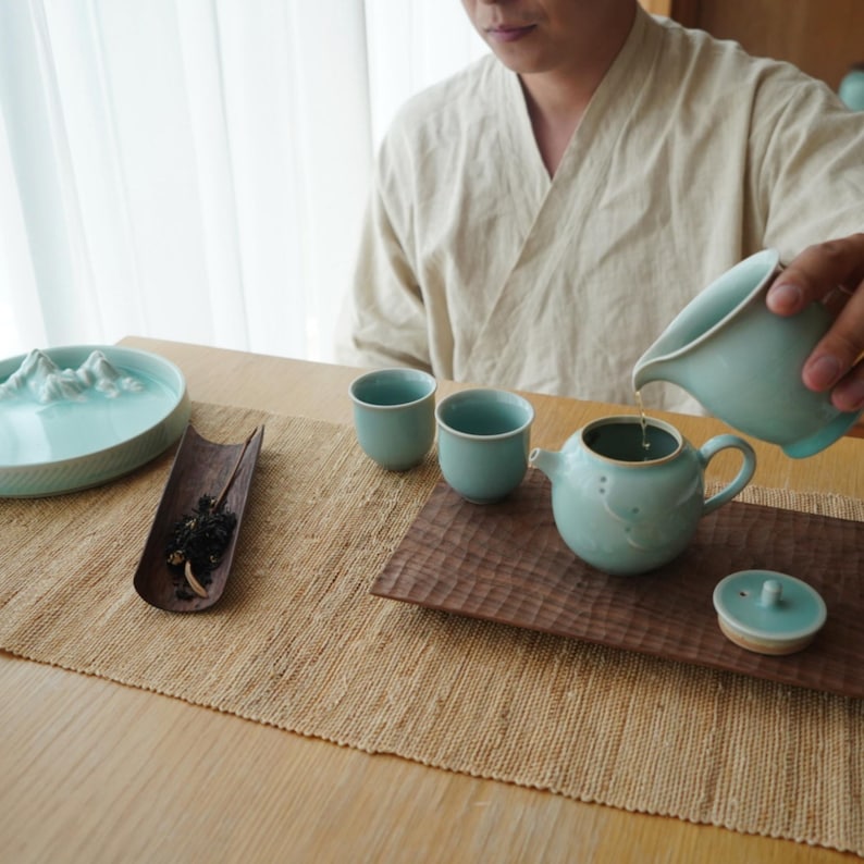 May include: A tea ceremony setup with a light blue ceramic teapot, two cups, and a serving pitcher. The tea set is on a wooden tray and a woven mat. A person is pouring tea into the teapot. The scene evokes a sense of calm.