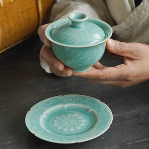 May include: A turquoise ceramic tea set with a floral pattern. The set includes a lidded bowl and a matching saucer. The bowl is held in a person's hands. The saucer is on a dark surface.