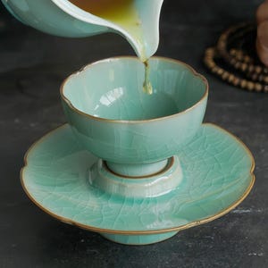 May include: A light mint green ceramic tea cup and saucer set with a visible crackle glaze finish and gold-toned edges. The cup sits on a matching saucer with a raised center, while golden tea is being poured into the cup from a pitcher.