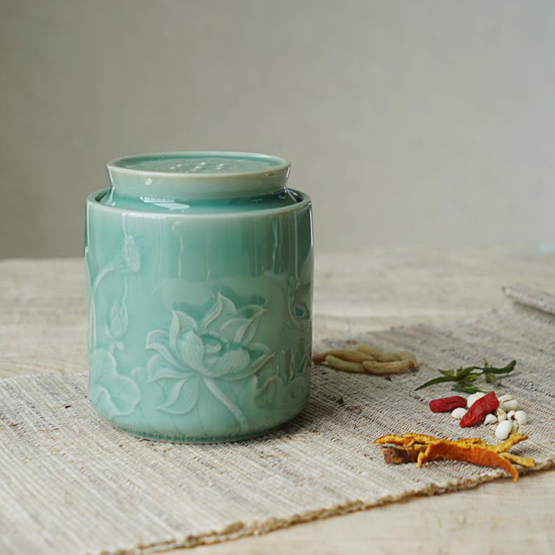 May include: A light teal ceramic jar with a lid, featuring a detailed embossed lotus flower design. The jar is cylindrical and sits on a neutral-toned surface. Dried herbs and spices are scattered around the jar.