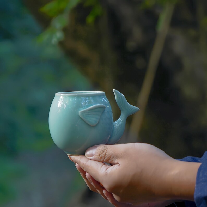 May include: A light blue ceramic mug shaped like a whale. The mug has a rounded body, a fin on the side, and a tail forming the handle. The mug is held in a person's hands against a blurred green background.