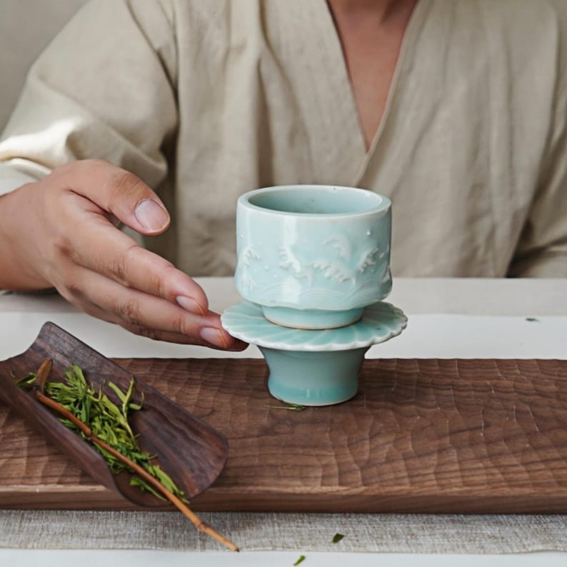May include: A light blue ceramic tea cup and saucer set with a floral design. The cup is cylindrical with a detailed relief pattern. The saucer has a scalloped edge. The set is displayed on a wooden tray with tea leaves.