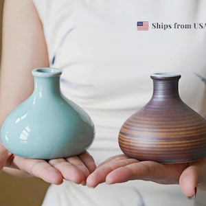 May include: Two small ceramic vases. One is a light teal colour, and the other is brown with horizontal lines. Both have a rounded body and a narrow neck. The vases are held in open hands.