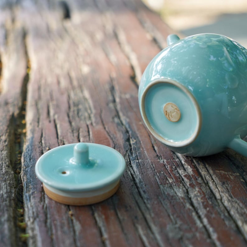 May include: A light blue ceramic teapot with a matching lid. The lid is off to the side, revealing a small hole. The teapot has a rounded shape and a small spout. The base of the teapot has a small, circular design.