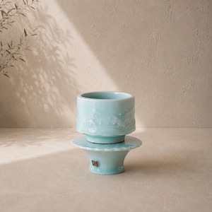May include: A light blue ceramic cup and saucer set. The cup has a textured wave design around the top. The saucer has a scalloped edge. The set is on a neutral background with shadows.