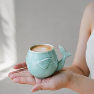 May include: A light blue ceramic mug shaped like a whale, with a tail handle and fin details. The mug is filled with a latte, featuring a decorative foam design. The mug is held in a person's hand.