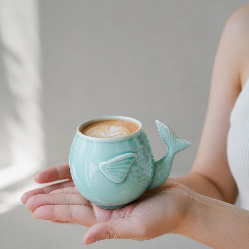 May include: A light blue ceramic mug shaped like a whale, with a tail handle and fin details. The mug is filled with a latte, featuring a decorative foam design. The mug is held in a person's hand.