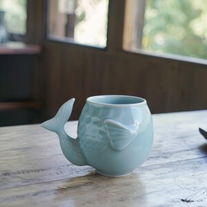 May include: A light blue ceramic mug shaped like a whale, with a tail handle and a fin on the side. The mug has a textured surface and sits on a wooden table. The mug is designed for beverages.